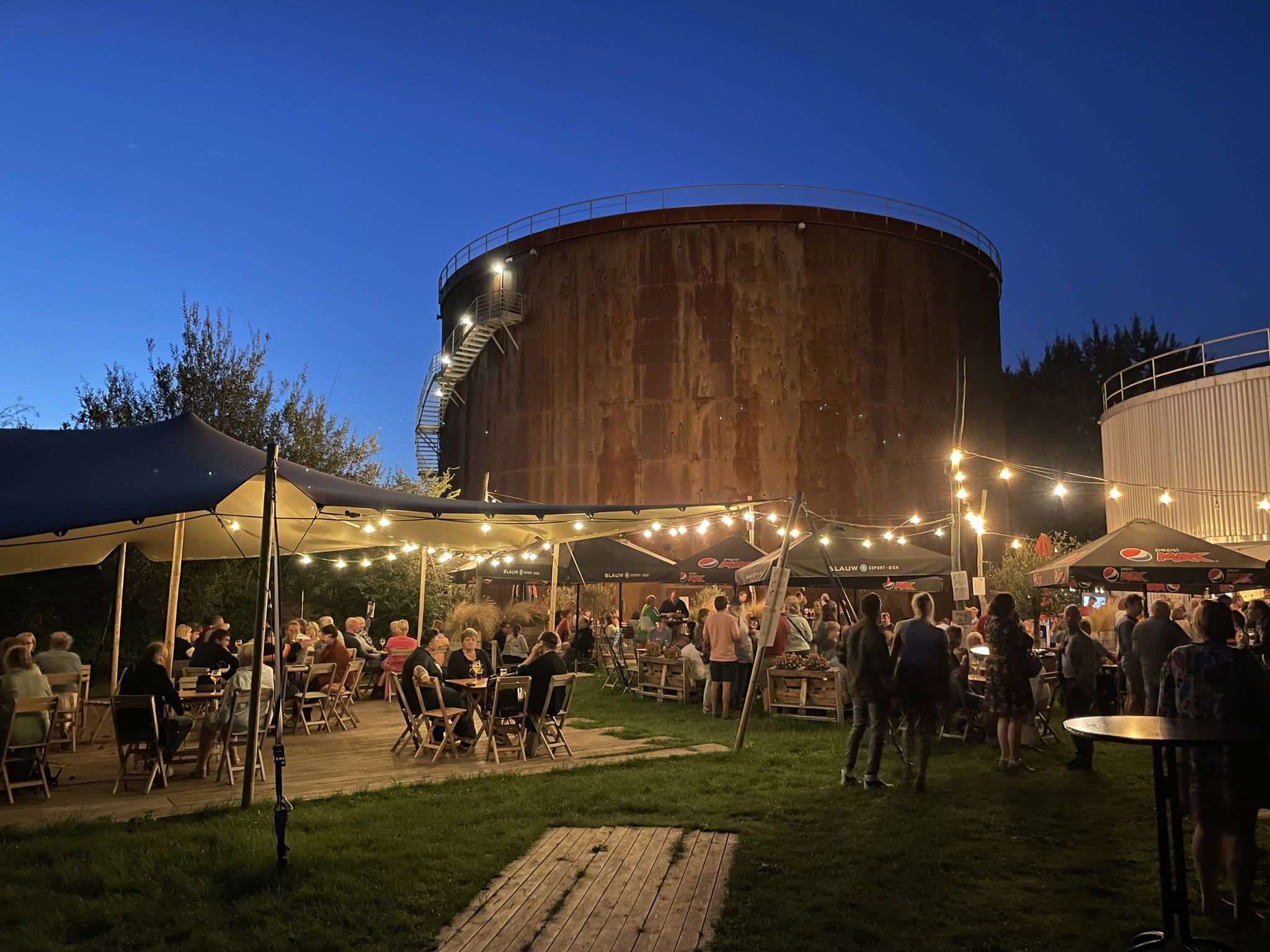 Het zomerterras van Bar Elektriek in het blauwe uur, aan de voet van de verlichte tank
